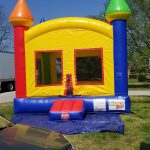 A colorful inflatable bounce house with red, yellow, green, and blue sections sits on grass next to a parked green car under a clear blue sky.