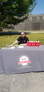 A man with a beard and sunglasses sits at an outdoor table covered with a gray tablecloth labeled "AAWDC Community Career Connections." Red water bottles and brochures are displayed on the table.