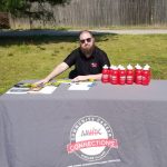 A man with a beard and sunglasses sits at an outdoor table covered with a gray tablecloth labeled "AAWDC Community Career Connections." Red water bottles and brochures are displayed on the table.