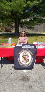 A woman with curly hair and sunglasses sits at a table covered with brochures and flyers. The table has a cloth displaying the Anne Arundel County Department of Aging & Disabilities seal. Trees and a fence are in the background.