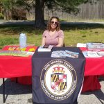 A woman with curly hair and sunglasses sits at a table covered with brochures and flyers. The table has a cloth displaying the Anne Arundel County Department of Aging & Disabilities seal. Trees and a fence are in the background.