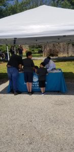A man and two children stand at a booth covered by a white canopy, interacting with a woman seated behind a table with a blue tablecloth that reads "Anacostia Neighborhood Library.