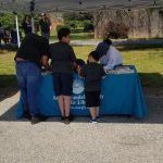 A man and two children stand at a booth covered by a white canopy, interacting with a woman seated behind a table with a blue tablecloth that reads "Anacostia Neighborhood Library.