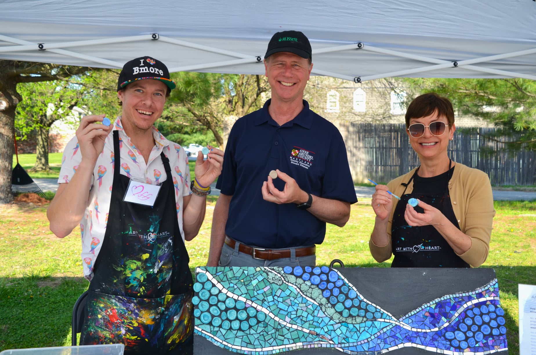 Three smiling adults stand under a white canopy, holding small mosaic pieces. The man on the left wears a colorful apron and “I ❤️ Bmore” hat, while the other two hold paintbrushes behind a wavy blue mosaic artwork.