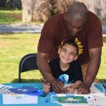An adult helps a smiling young boy draw on a tote bag at an outdoor arts and crafts table, surrounded by paper, markers, and craft supplies.