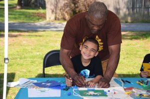 An adult helps a smiling young boy draw on a tote bag at an outdoor arts and crafts table, surrounded by paper, markers, and craft supplies.