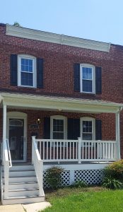 A two-story red brick house with white trim, black shutters, and a covered front porch with white railings and steps. There are bushes and green grass in the front yard.