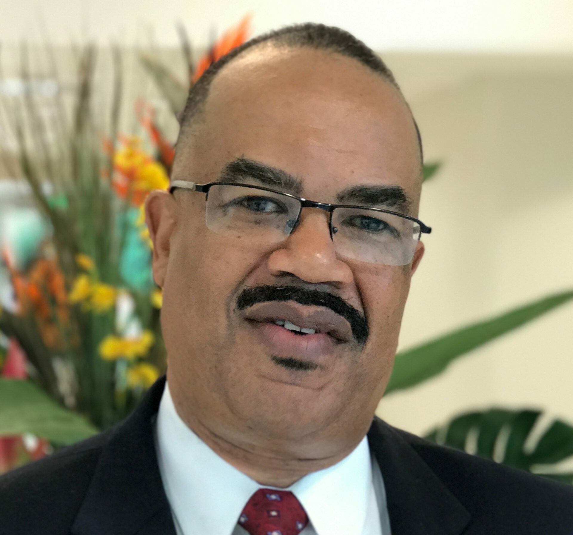A man wearing glasses, a dark suit, white shirt, and red tie stands indoors. He has a mustache and short hair. Colorful flowers and green leaves are visible in the blurred background.