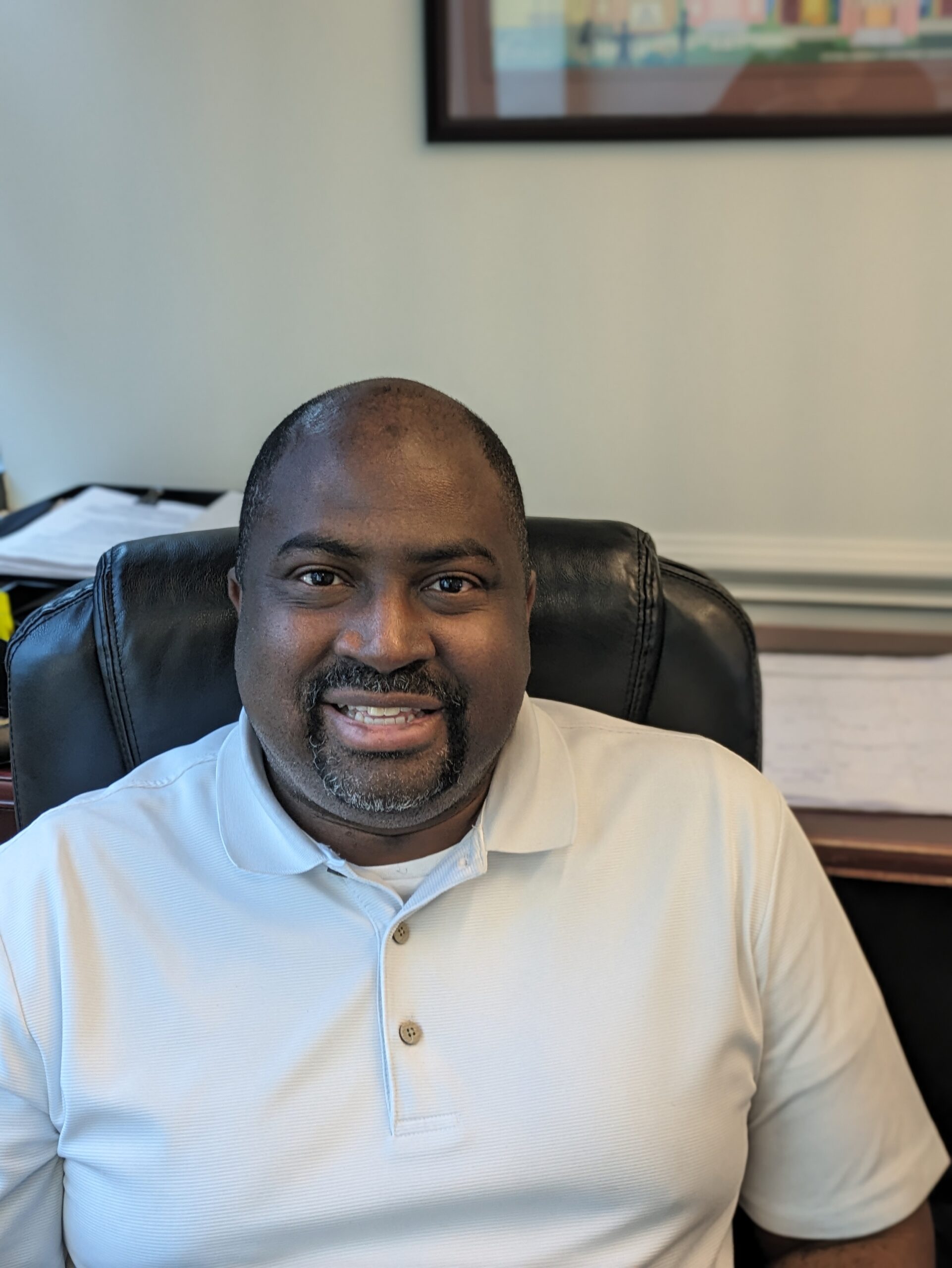 A man with short hair and a goatee, wearing a white polo shirt, sits in a black office chair and smiles at the camera. Office papers and a framed picture are visible in the background.
