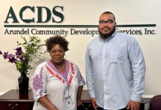 Two people stand and smile in front of a wall sign that reads "Arundel Community Development Services, Inc." There are flowers on the left and a desk in the office setting.