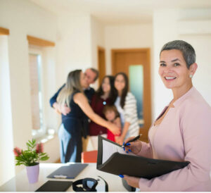 A smiling real estate agent stands holding a clipboard in a bright office while a happy family hugs in the background, celebrating together.